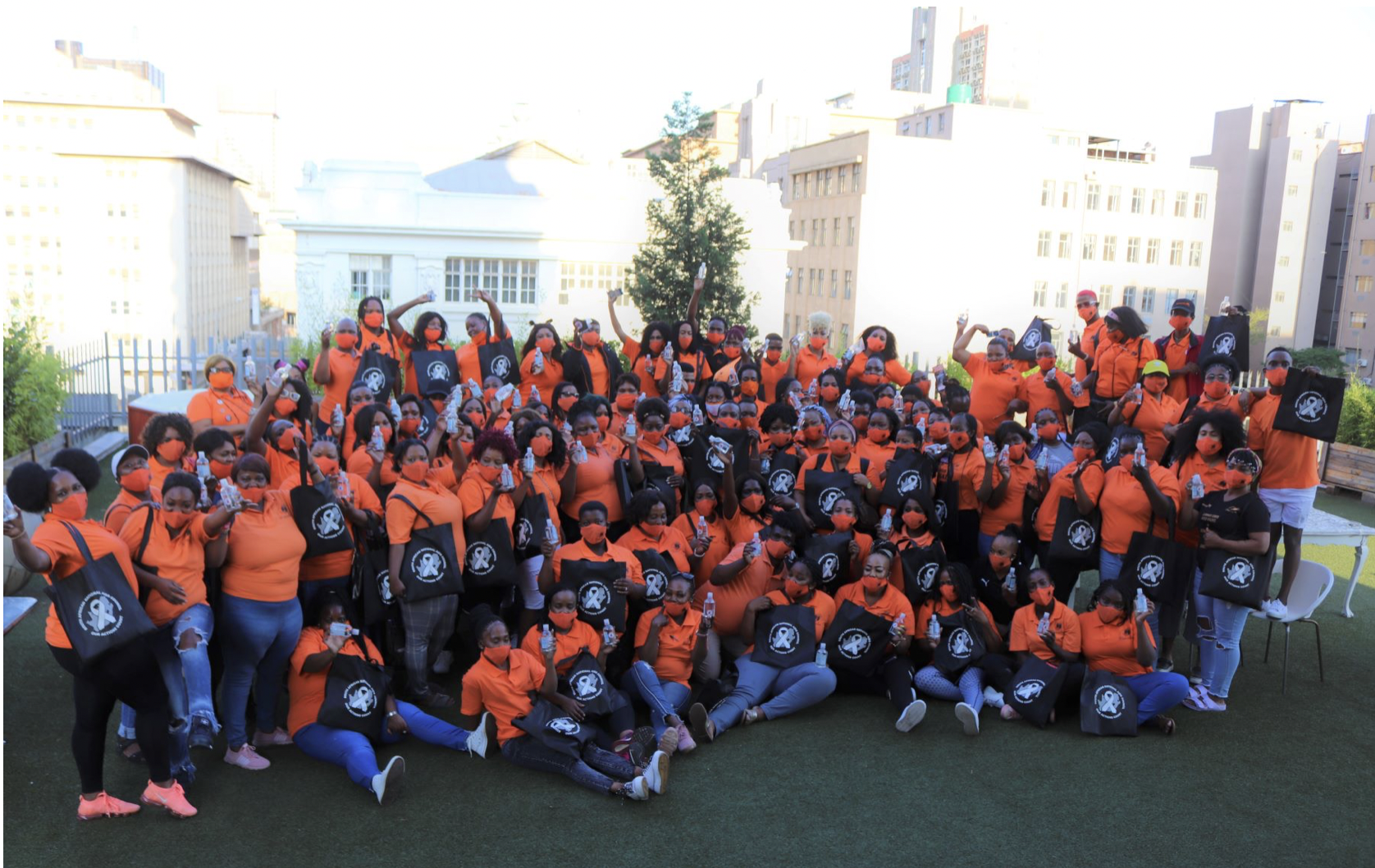 A group photo of about 90 people at a 2020 national Sisonke meeting. They are all wearing bright orange shirts and holding black tote bags. They are all wearing face masks.