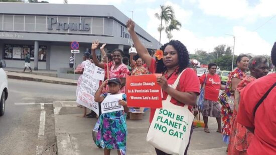 image of a climate justice rally. Sign reads: Climate Change Is a Feminist Issue - women Let's Act! Activist is wearing a bag that reads: Sori Be...Planet B Ino Gat.
