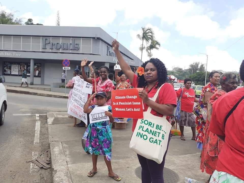 image of a climate justice rally. Sign reads: Climate Change Is a Feminist Issue - women Let's Act! Activist is wearing a bag that reads: Sori Be...Planet B Ino Gat.