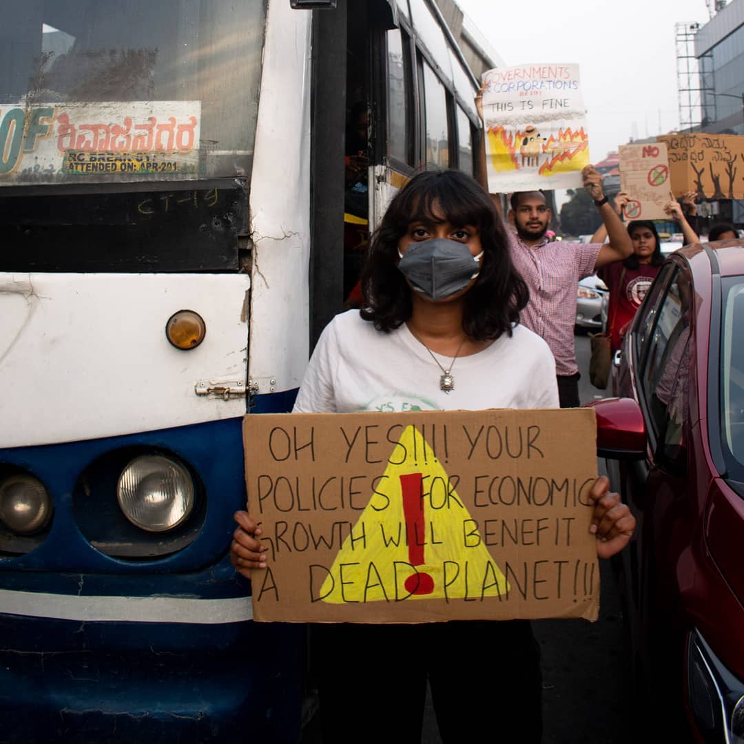 Disha Ravi holding a sign at a rally that reads "Oh Yes!!! Your Policies For Economic Growth Will Benefit a Dead Planet!!!