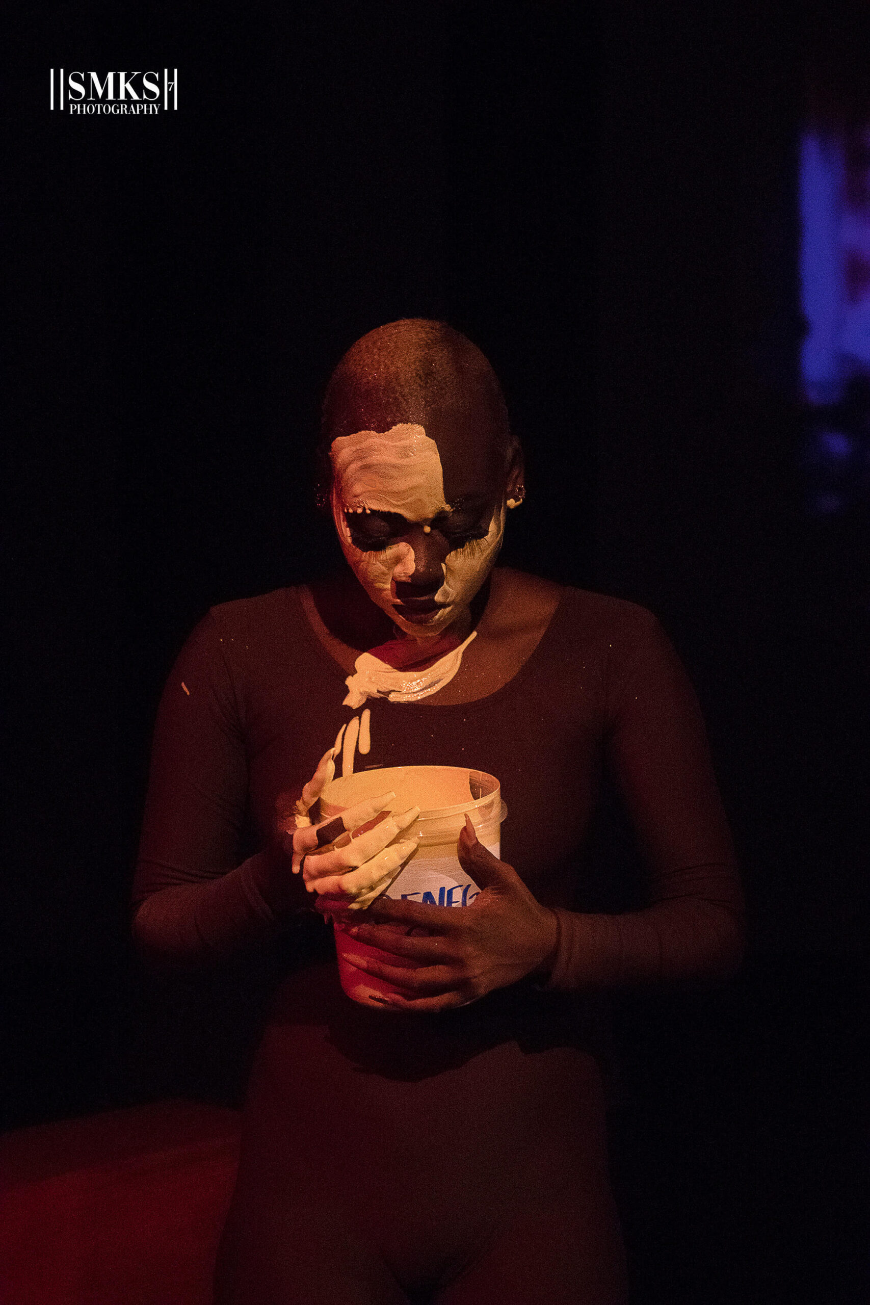 A Black figure in a shadow looks downward. She is holding a bucket of white paint and has smeared white paint on most of her face.