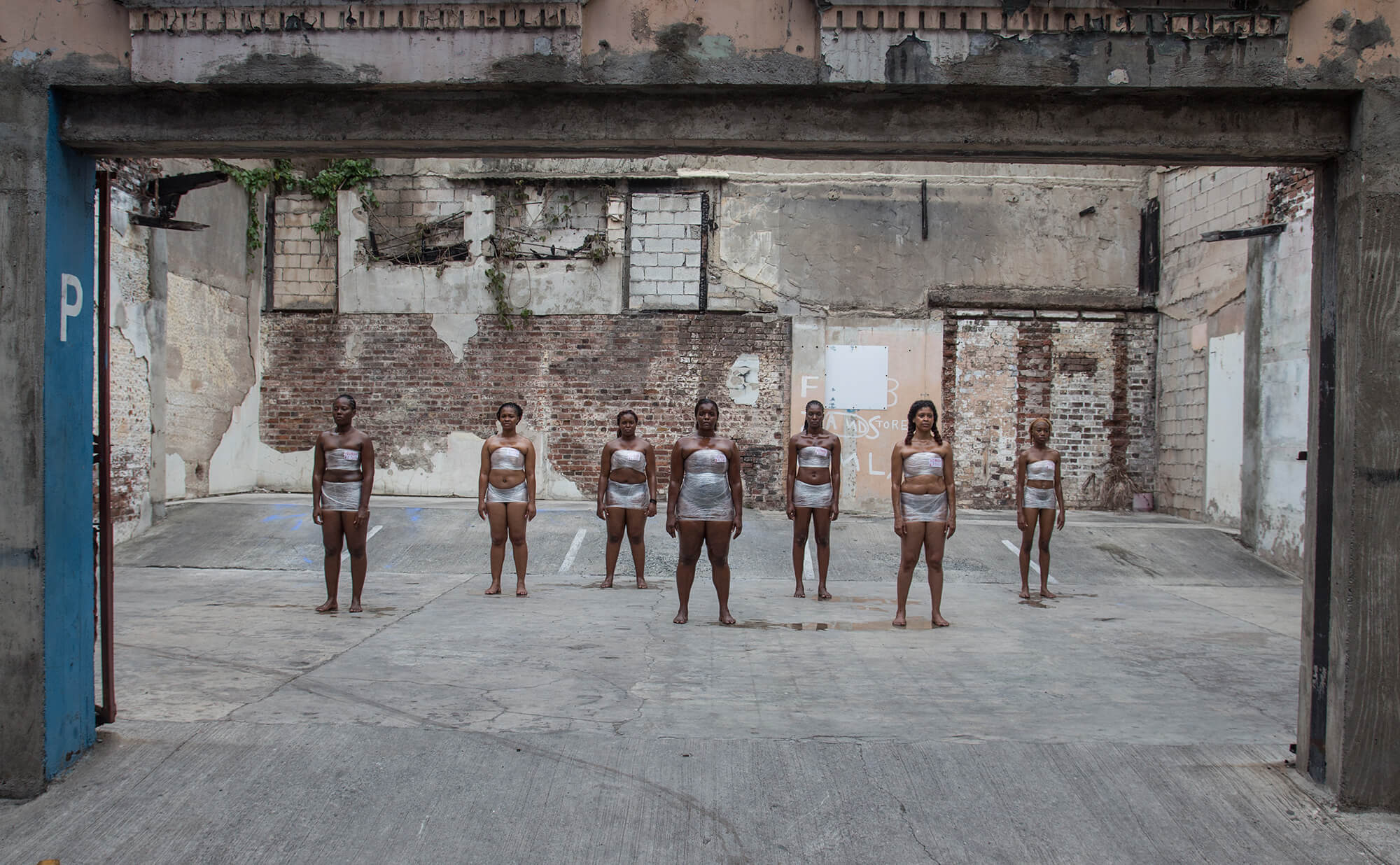 A group of seven Black women stand in a concrete space. They all wear plastic wrap. The photo is documentation of “Woman for Sale,” an experimental performance installation. The performance intended to physicalize the "meat market" of women as inanimate objects, priced according to the cultural beauty standards surrounding complexion, age, weight, hair texture, and shape.