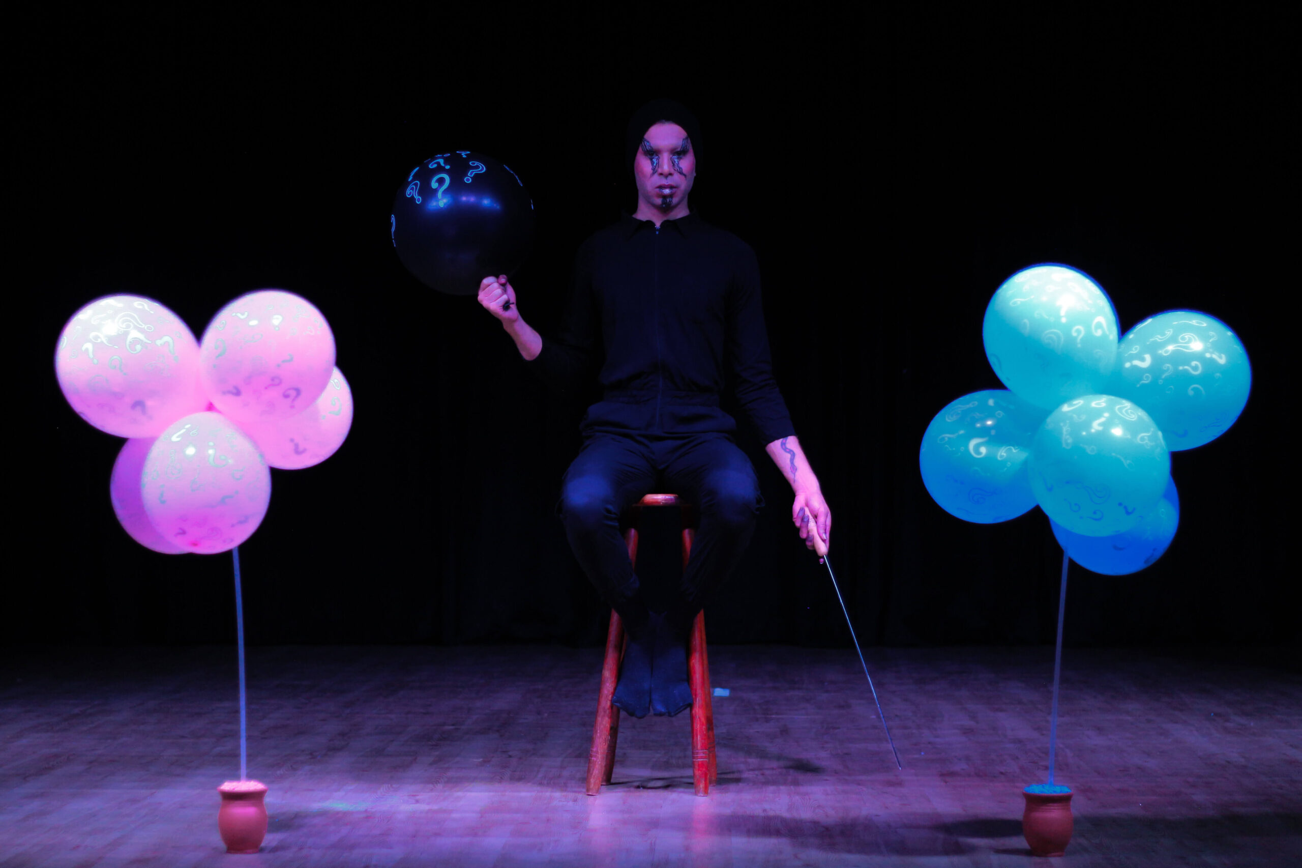 Jono Lena sits with a balloon on one hand and a long, pointy stick in the other. Pink balloons are placed on one side of her and blue balloons on the other.