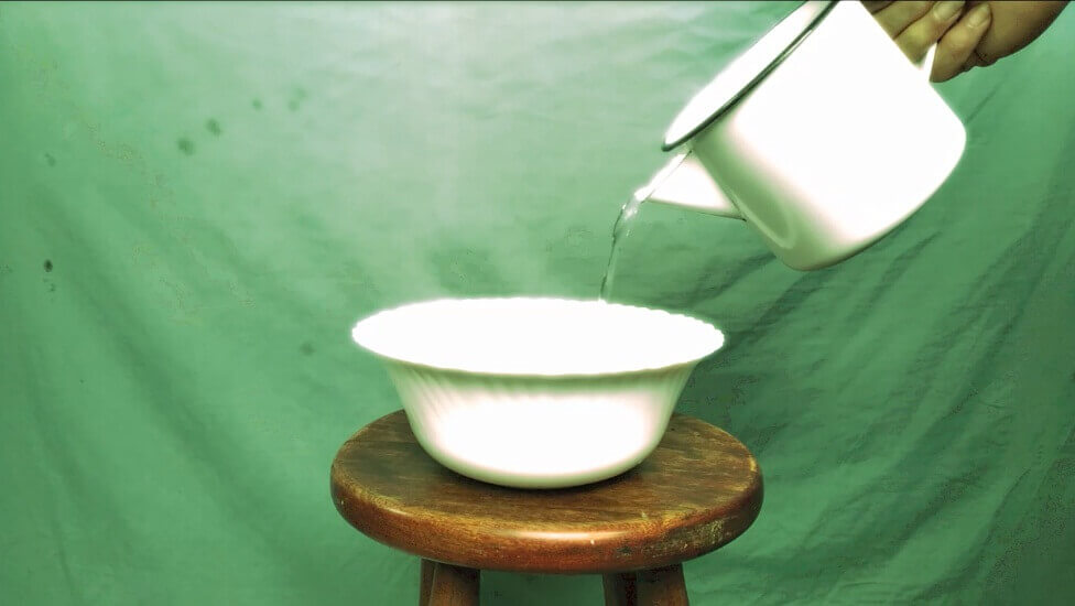 A hand pours a clear liquid from a white pitcher into a white basin. The background is bright green.