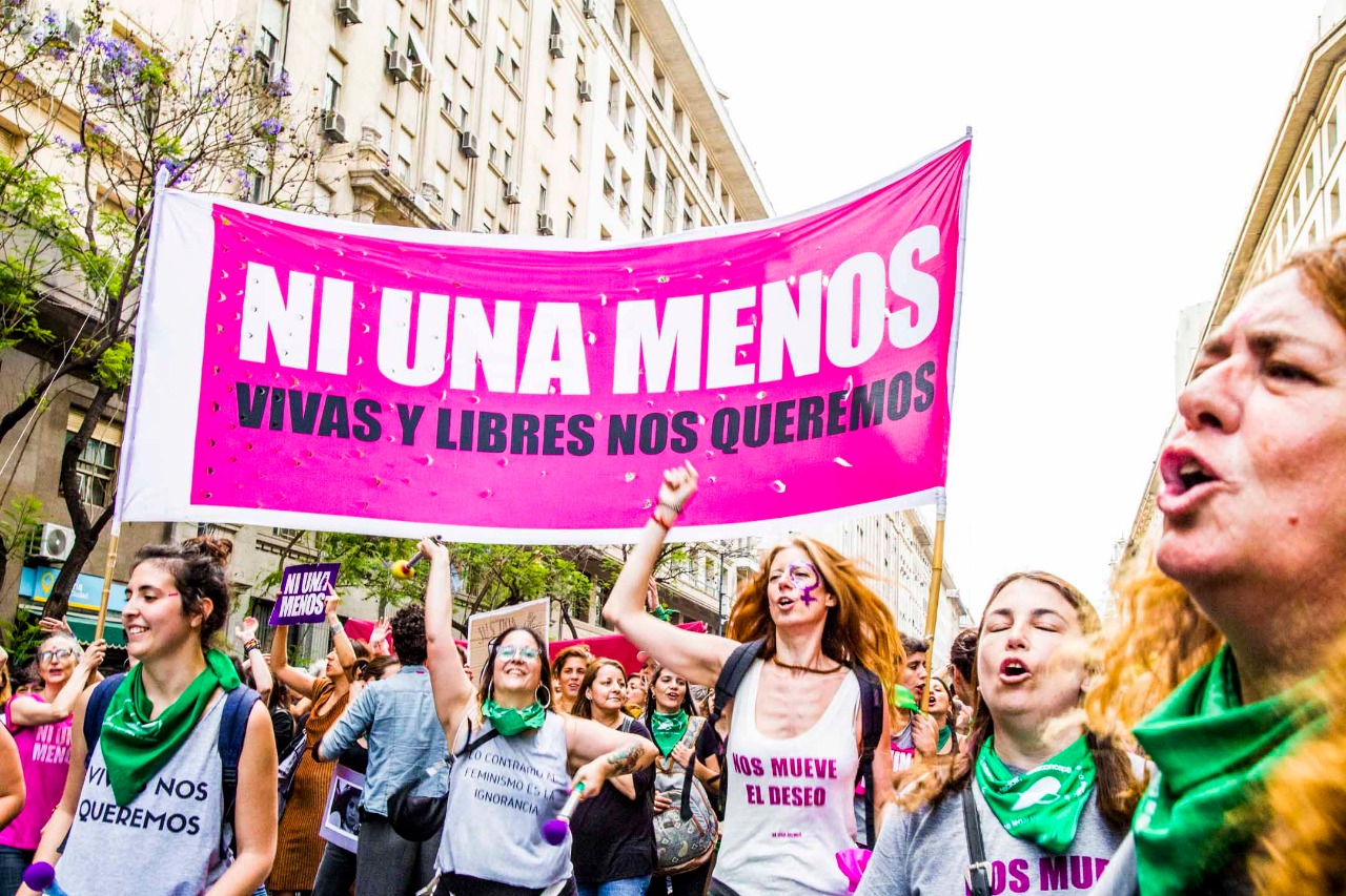 group of activists at a protest, people in the foreground hold up a large pink sign that reads "NI UNA MENOS VIVAS Y LIBRES NOS QUEREMOS"