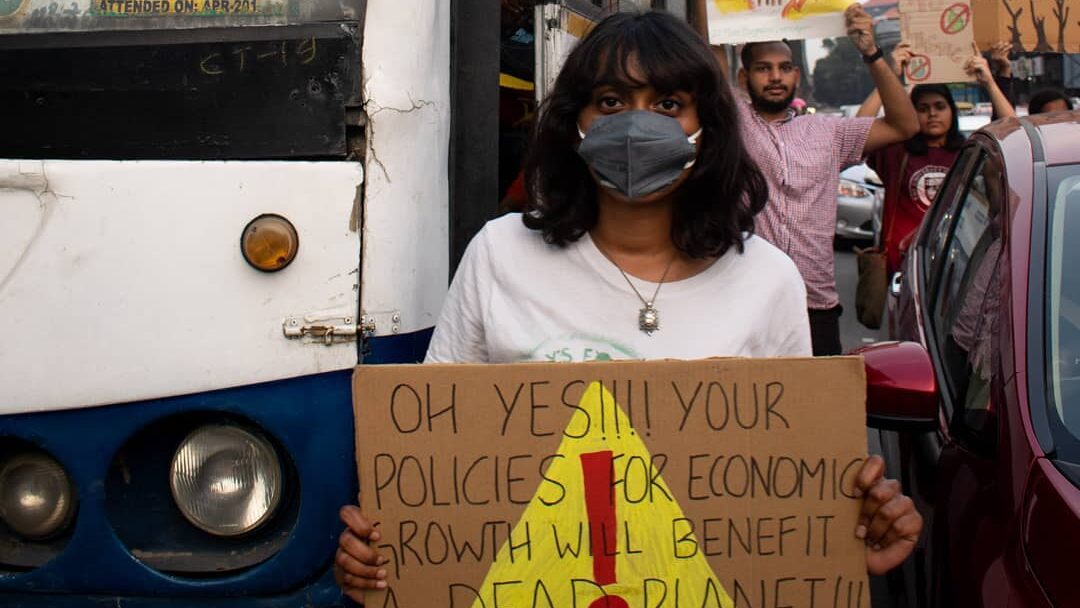 Disha Ravi, a feminist, climate, and environmental activist holds up a cardboard sign that reads "OH YES!!!! YOUR POLICIES FOR ECONOMIC GROWTH WILL BENEFIT A DEAD PLANET!!!"
