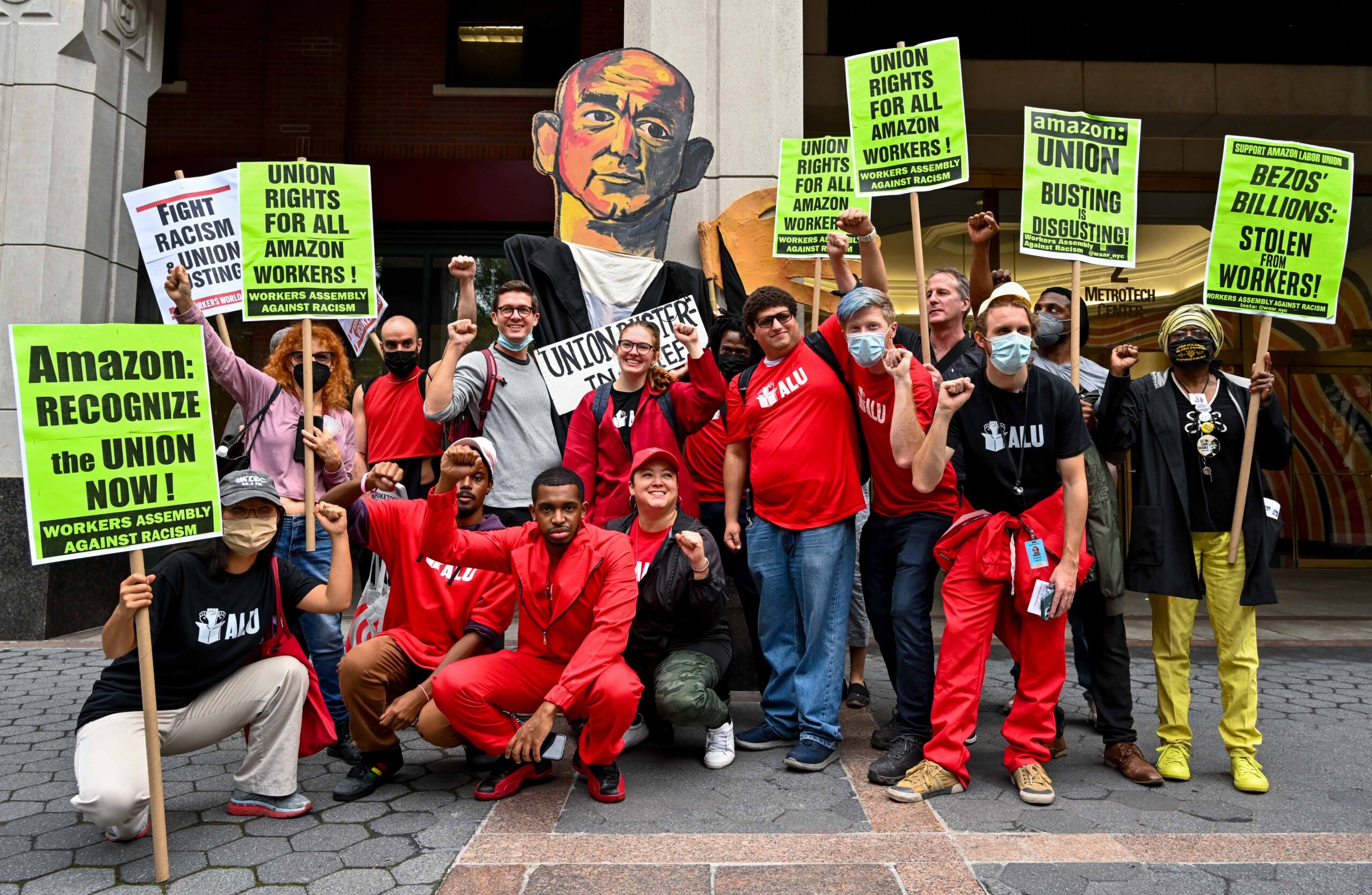 A group of Amazon warehouse workers outside the National Labor Relations Board. They are wearing mostly red, holding neon green protest signs, and raising up their fists. An illustrated giant poster of Jeff Bezos sits behind them.