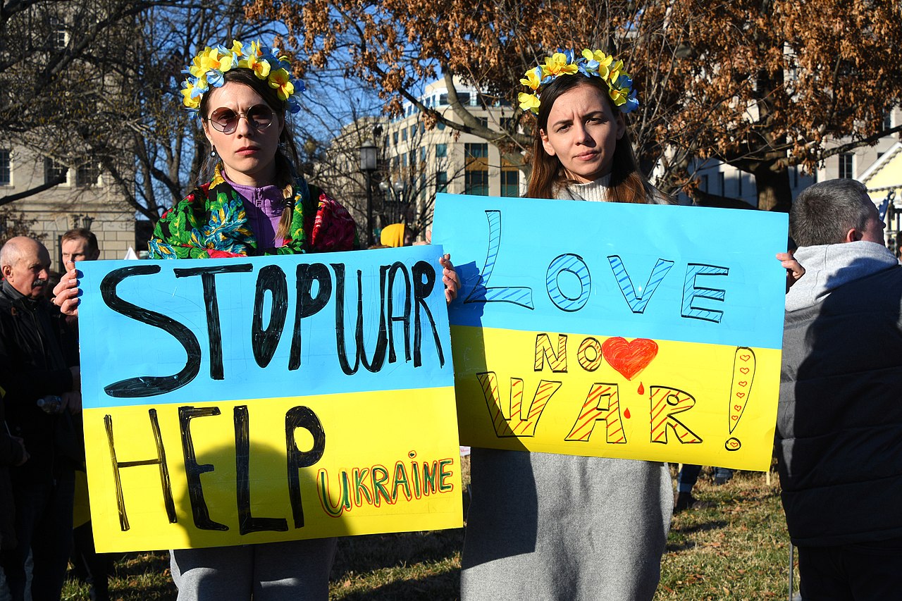 Photo from a protest against the war in Ukraine where two people wearing floral crowns in yellow and blue hold up two large blue and yellow protest signs that say "STOP WAR HELP UKRAINE" and "LOVE NO WAR"