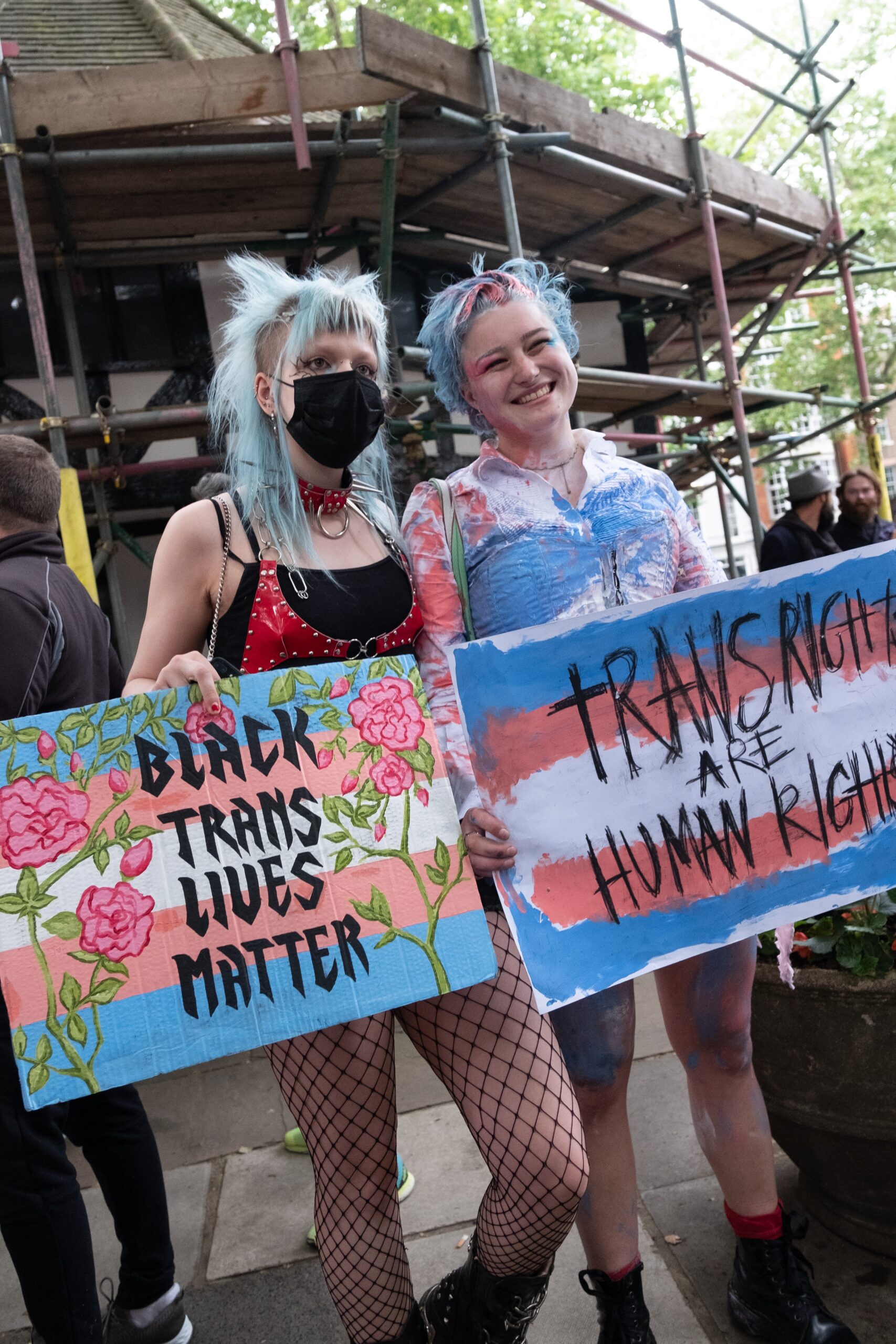 Two protestors stand with light blue, pink, and white striped signs that read "BLACK TRANS LIVES MATTER" and "TRANS RIGHTS ARE HUMAN RIGHTS". One is wearing a mask and the other is smiling.