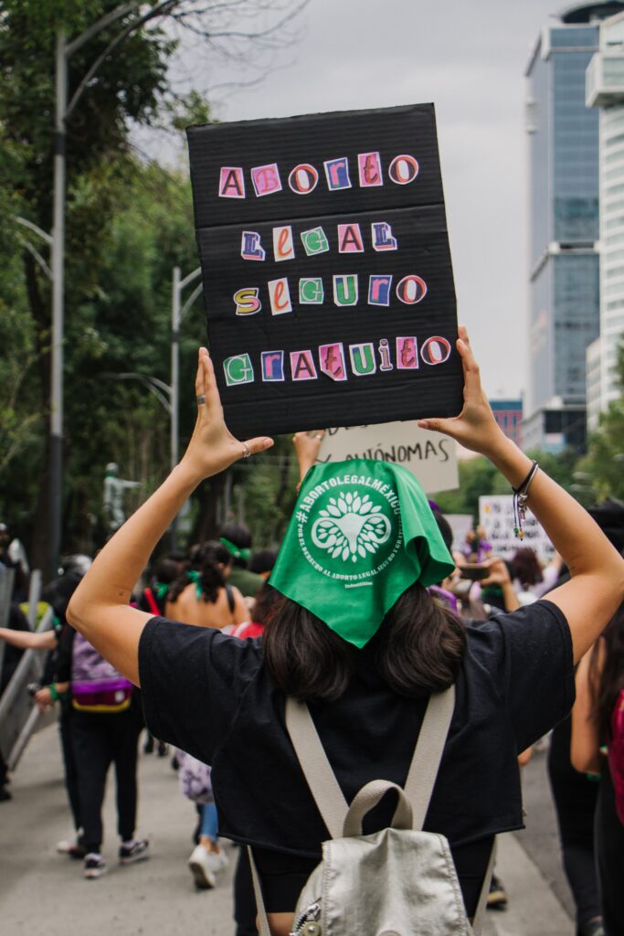 A protestor with a green hankerchief on her head holds up a sign with cut out letters that reads "Aborto legal seguro gratuito"