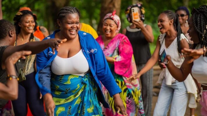 Members of the Movement Committee dance during a visit to a local indigenous community in Kribi, Cameroon. Caryn, in blue, is in the forefront, with Joséphine smiling on the left, Bernadette smiling in pink, and Aichatou and Nafissate on the right. In the background, Prisca films on her cell phone in the back next to Hermine.