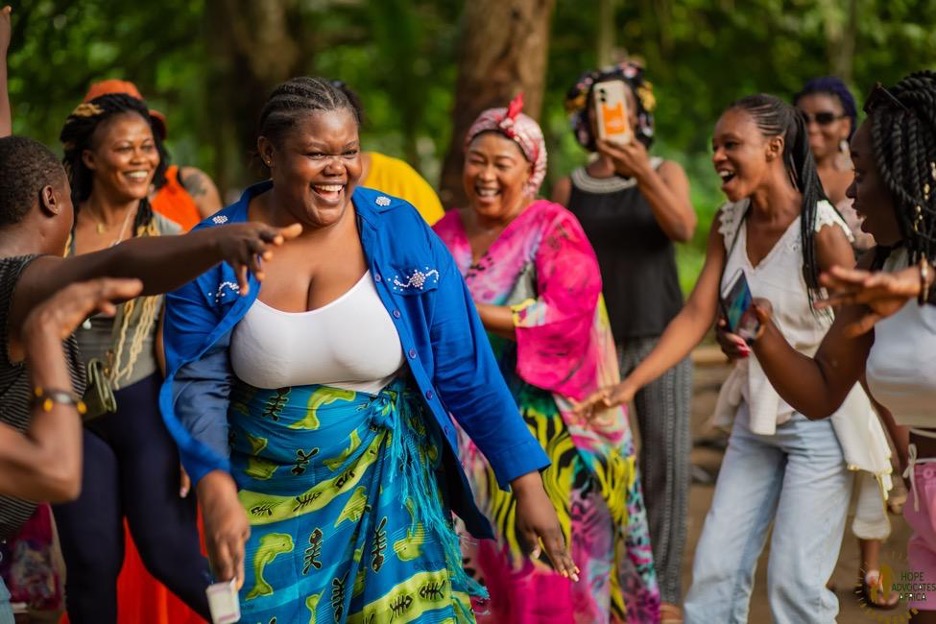 Members of the Movement Committee dance during a visit to a local indigenous community in Kribi, Cameroon. Caryn, in blue, is in the forefront, with Joséphine smiling on the left, Bernadette smiling in pink, and Aichatou and Nafissate on the right. In the background, Prisca films on her cell phone in the back next to Hermine.